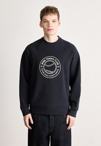 Tumši zils džemperis ar ribotu aproci un apakšu, ar apaļu baltu izšuvumu, kurā rakstīts "BOSS TENNIS CLUB 1997".