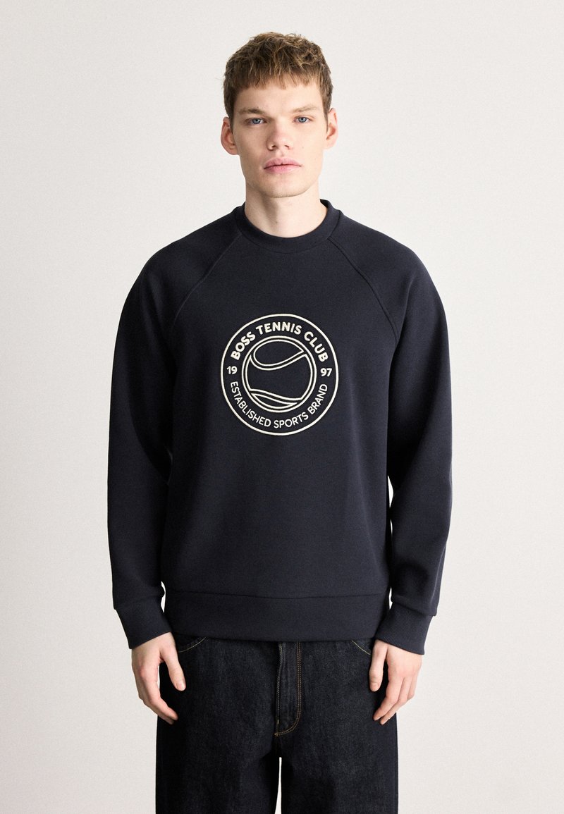 Tumši zils džemperis ar ribotu aproci un apakšu, ar apaļu baltu izšuvumu, kurā rakstīts "BOSS TENNIS CLUB 1997".
