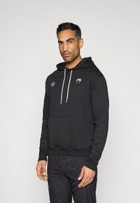 Sweatshirt preta com capuz feita de algodão, com bolso frontal, cordões brancos e detalhes de logotipo no peito. Textura lisa.