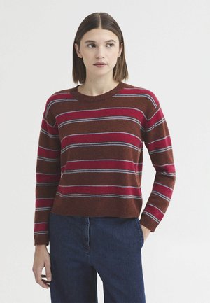 Femme aux cheveux bruns et raides portant un pull rayé marron et rouge et un jean bleu foncé, debout avec une main dans la poche.