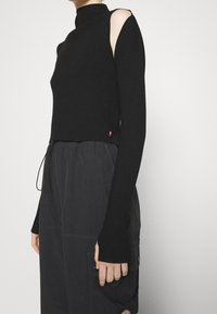 Schwarzes Strickoberteil mit hohem Kragen und schulterfreiem Ausschnitt. Es verfügt über lange Ärmel und ein cropped Design, ergänzt durch ein kleines Logo.