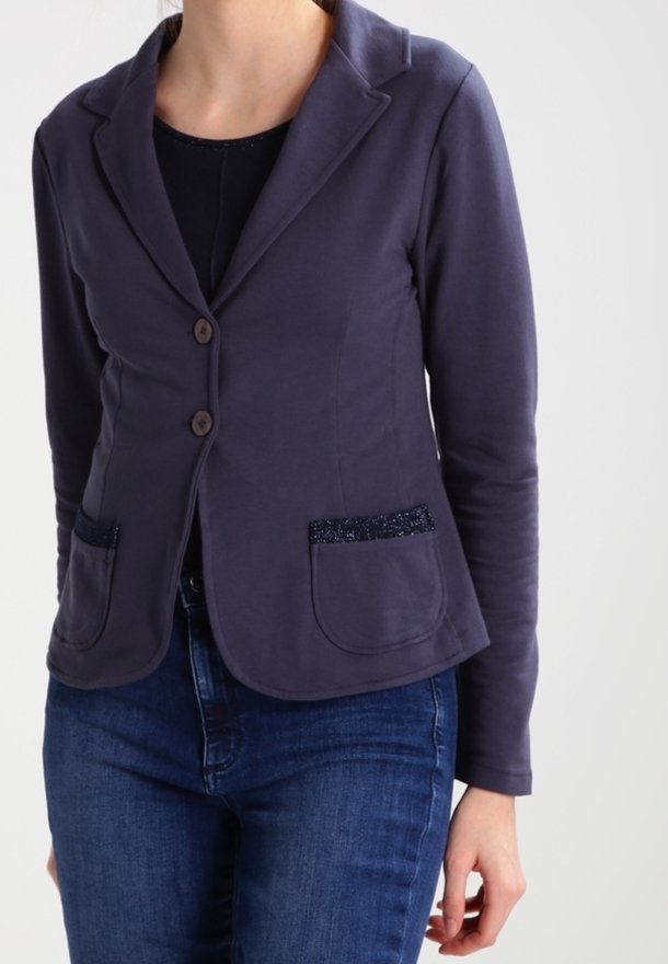 Femme portant un blazer bleu marine cintré avec deux boutons et des poches plaquées, par-dessus un haut noir et un jean bleu, debout devant un fond uni.