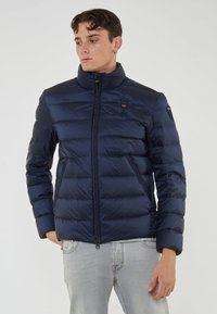 Uomo che indossa un giubbotto trapuntato blu scuro con tasche con zip e jeans chiari, che guarda di lato su uno sfondo neutro.