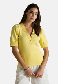 Naine kannab kollast lühikeste varrukatega pluusi, millel on nupud, ja valgeid pitsilisi pükse, poseerides käed rinna ees kokku surutud.