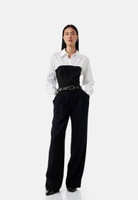 Zwarte fitted top met een witte blouse eronder, cinched met een zwarte riem. Wijde zwarte broek, gladde stof, en zwarte puntige schoenen.