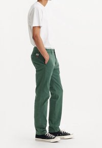Pantalons en coton verts avec une coupe slim, poches latérales et un patch logo rouge sur la taille. Associés à un t-shirt blanc uni et des baskets noires.