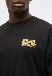 Μαύρο μπλουζάκι με κίτρινο κείμενο "We Are Juventus" στο στήθος, φορεμένο από άτομο με σκοτεινό δέρμα, ορατό από τα χείλη μέχρι το πάνω μέρος του στήθους.