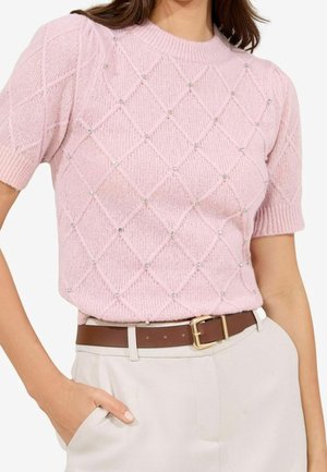 Jasnoróżowy, dziergany sweter z krótkim rękawem w diamentowy wzór, zdobiony kryształkami, zestawiony z brązowym paskiem na jasnobeżowych spodniach.