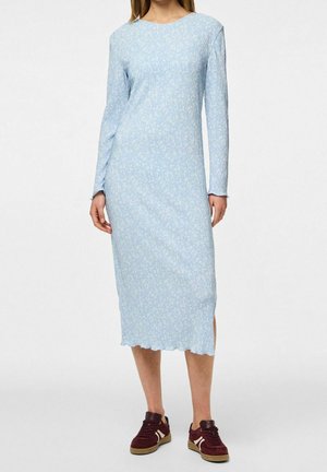 Kobieta w błękitnej, długorękawowej sukience midi z subtelnym wzorem, połączonej z bordowymi trampkami na białym tle.