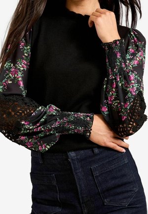 Vrouw draagt een zwarte top met bloemenprint en geplooide mouwen met geplooide manchetten en zwarte kanten inzetstukken, gecombineerd met donkere, hooggesneden spijkerbroek.