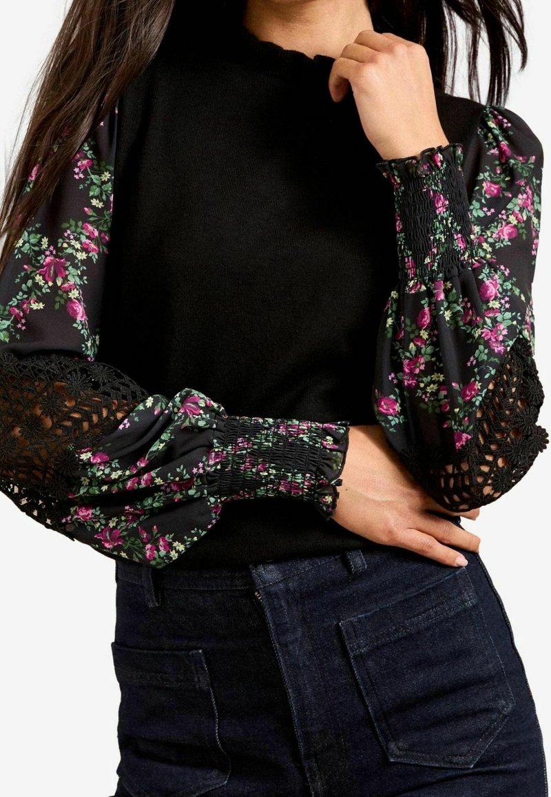 Femme portant un haut noir avec des manches bouffantes à fleurs, dotées de poignets froncés et d'inserts en dentelle noire, associées à un jean foncé taille haute.