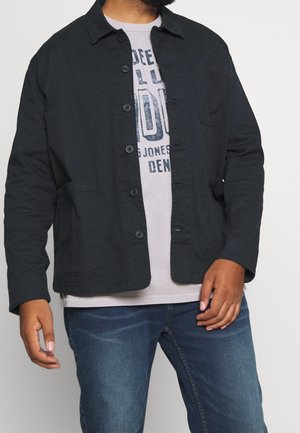 Mand iført en sort jakke med knapper over en lys t-shirt med grafik og blå jeans, stående mod en ensfarvet baggrund.