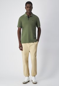 Grünes Poloshirt mit weißem Besatz, beiger tapered Hose und weißen Sneakern. Das Shirt hat ein kleines Logo auf der Brust.