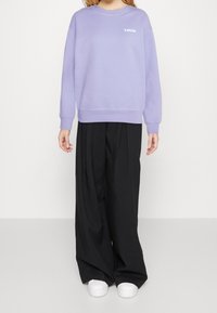 Sudadera lila clara con logo blanco, cuello redondo y mangas largas, combinada con pantalones negros holgados y zapatillas blancas.