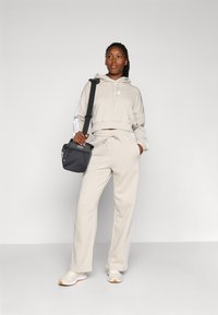 Beige cropped hoodie with drawstrings and white side stripes, paired with matching beige sweatpants and white sneakers. Holds a black bag.