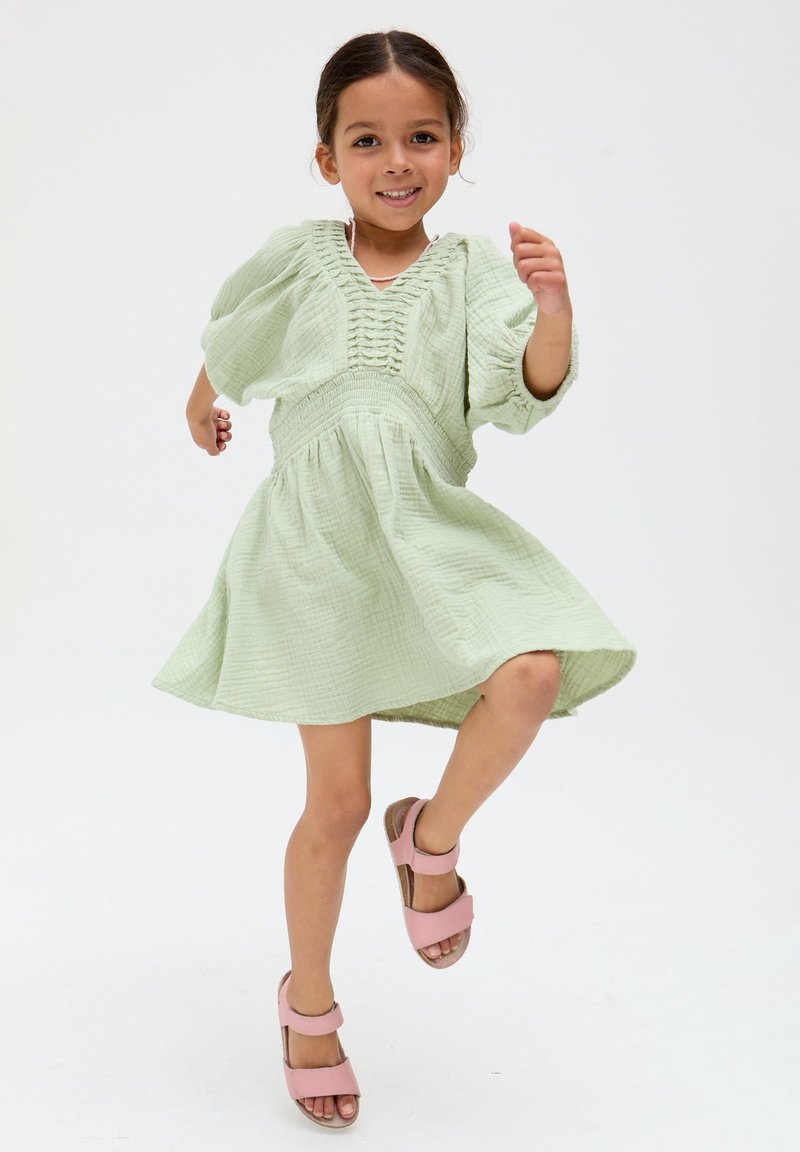 Junges Mädchen lächelt und tanzt, trägt ein hellgrünes Kleid mit Puffärmeln und pinke Sandalen vor weißem Hintergrund.