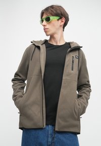 Καφέ φλις ζακέτα με κουκούλα, μπροστινές τσέπες με φερμουάρ, και μαύρη μπλούζα από κάτω. Φορώντας με μπλε τζιν και πράσινα γυαλιά ηλίου.