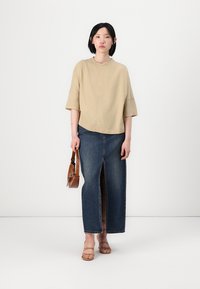 Beige short-sleeve top paired with a long denim skirt featuring a front slit. Model holds a brown handbag and wears strappy sandals.