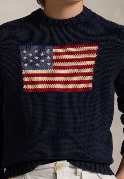 Maglione a maglia blu navy con il disegno della bandiera americana in rosso, bianco e blu lavorato a maglia sul petto, indossato con pantaloni chiari.