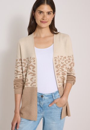 Femme portant un cardigan beige avec imprimé léopard sur les manches et devant, un top blanc et un jean bleu clair, une main dans la poche.