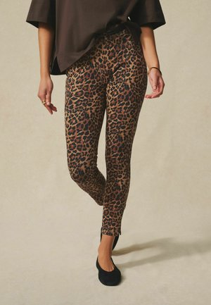 Woman wearing brown and black leopard print skinny pants, black flats, and a loose black top, standing on a neutral background.