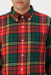Camicia a quadri rossa, verde e gialla con colletto, dotata di bottoni bianchi e taschino sul petto. Tessuto di texture.