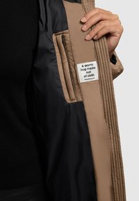 Beige ja must padded jakk, mille siseküljel on silt tekstiga, küljetasku ja tekstuuriga õmblus äärealadel.