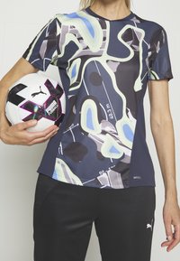 Persoon draagt een sportshirt van Puma met abstract patroon en houdt een witte en zwarte Puma voetbal ter hoogte van de taille vast.