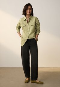 Hellgrünes Hemd mit Knöpfen und zwei Brusttaschen, kombiniert mit hoch taillierter schwarzer Jeans und olivgrünen Ballerinas. Weicher Stoff, lässige Passform.