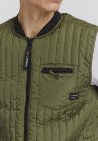 Chaleco acolchado verde oliva con cremallera negra, cuello de canalé y un bolsillo en el pecho con cremallera que presenta la etiqueta de la marca. Textura de tela suave.