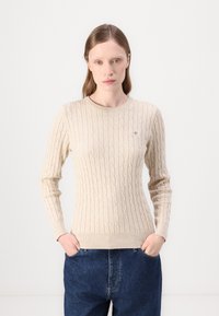 Pulover bej din tricotaj cu cablu, cu mâneci lungi și guler rotund. Dispune de un tiv și mansete ribbed, cu un mic logo brodat pe piept.