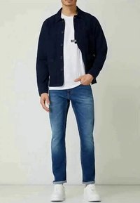 Hombre vistiendo una chaqueta negra de botones, camiseta blanca, jeans azules enrollados en los tobillos y zapatillas blancas, de pie contra un fondo liso.