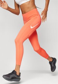 Oranžové sportovní legíny s vysokým pasem, s bílým logem Nike na boku, doplněné černými sportovními botami.