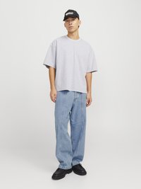 Camisa de algodón gris de gran tamaño, corte relajado, con mangas cortas. Pantalones vaqueros de pierna ancha en azul claro con detalles de costura en el frente. Gorra negra con logo y zapatos negros chunky.
