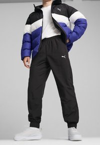 Modrá, černá a bílá péřovka se zipem; černé jogger kalhoty s elastickým pasem a logem; bílé slip-on boty s texturou.