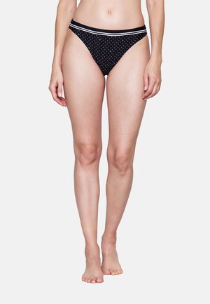 Frau trägt schwarze Bikinihose mit weißen Punkten und einem weiß gestreiften Bund, steht barfuß vor weißem Hintergrund.