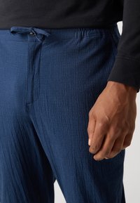 Marineblauwe textuur broek met een elastische tailleband en trekkoord, met een losse pasvorm en een subtiel ruitpatroon. Hand zichtbaar die de taille vasthoudt.