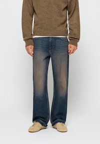 Tmavomodré džíny s rovnými nohavicami, so šúchanými detailmi, dizajnom na päť vreciek a svetlým prešívaním; skombinované s hnedým pleteným sveterom.