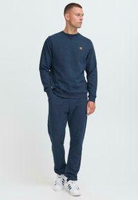 Férfi áll a zsebébe tett kezekkel, sötétkék pulóverben és azonos színű nadrágban, fehér sportcipőben, oldalra nézve.