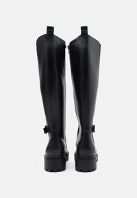 Hohe schwarze Lederstiefel mit robusten Sohlen, rückseitigem Reißverschluss und kleinen verstellbaren Riemen am Knöchel, von hinten betrachtet.