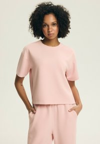 Kobieta o kręconych włosach, ubrana w luźny, bladoróżowy top z krótkim rękawem oraz pasujące spodnie, stoi z rękami w kieszeniach na jednolitym tle.