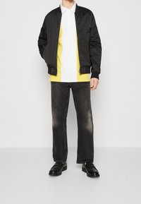 Black bomber jacket over a yellow and white striped polo shirt, paired with dark grey straight-leg jeans and black shoes.
