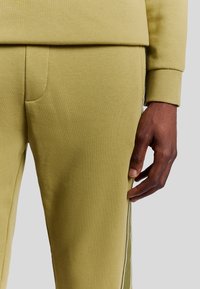 Olivgrüne Sweatpants aus einem weichen Baumwollmix mit schmaler Passform, ausgestattet mit Seiten Taschen und weißen Akzentstreifen entlang der Nähte.