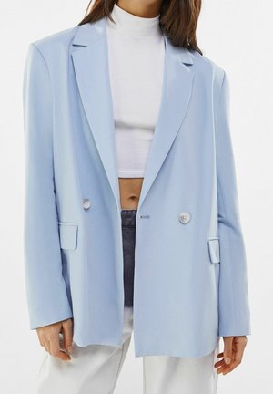 Vrouw die een pastelblauw dubbelrijig blazer draagt over een witte cropped coltrui en witte broek, staand tegen een effen achtergrond.