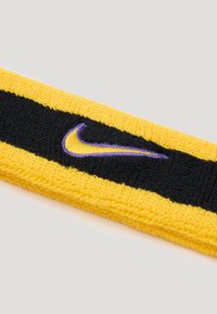 Nike sportska traka za glavu u žutoj boji s crnim središnjim prugom. Sadrži izvezeni ljubičasti Nike logo. Napravljena je od teksturiranog elastičnog pamuka.
