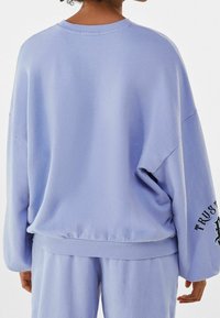 Sweatshirt de couleur violette claire, avec un col rond, une coupe oversize et un ourlet côtelé. La manche gauche affiche le texte "CONFIANCE" dans une couleur contrastante.