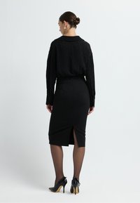 Schwarze Bluse mit strukturiertem Design, Kragen und langen Ärmeln; kombiniert mit einem schwarzen Bleistiftrock mit einem Schlitz hinten und hohen Schuhen.