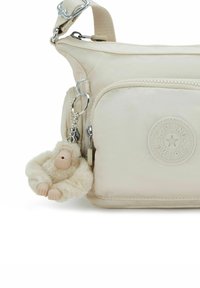 Beige Kipling Umhängetasche mit vorderer Reißverschlusstasche und angebrachtem, pelzigen Affen-Schlüsselanhänger in passendem Beigeton.