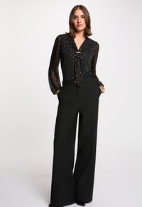 Blouse noire avec des pois métalliques, col noué et manches transparentes, associée à un pantalon large noir taille haute. Texture lisse sur l'ensemble.
