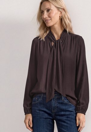Femme blonde portant un chemisier noir à manches longues avec un col noué et un jean bleu, souriante, sur un fond neutre uni.